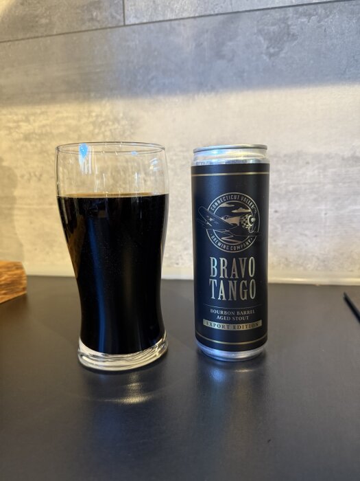 En glas med mörk dryck och en burk märkt "Bravo Tango, Bourbon Barrel Aged Stout" på ett bord.
