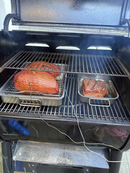Fläskkött röks i en grill, placerat i metallformar med termometrar, medan rök stiger uppåt under det öppna grillocket.