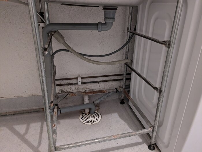 Under en diskbänk syns olika rörkopplingar, inklusive en vit frånluftsvärmepumpsrör och grå avloppsslangar till en tvättmaskin.