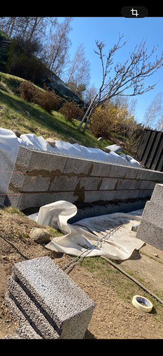 Mur av betongblock under konstruktion i en trädgård med grönska och träd, solig dag med verktyg och täckplast på marken.