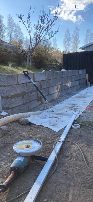 Mur av betongblock under uppbyggnad, omgivet av landskap och träd. Verktyg, som en vinkelslip, och skyffel ligger på marken.