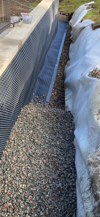 Dräneringssystem med svart plastnät mot en mur, kantad av vit presenning och grusfyllning samt en plastlåda med skruvar.