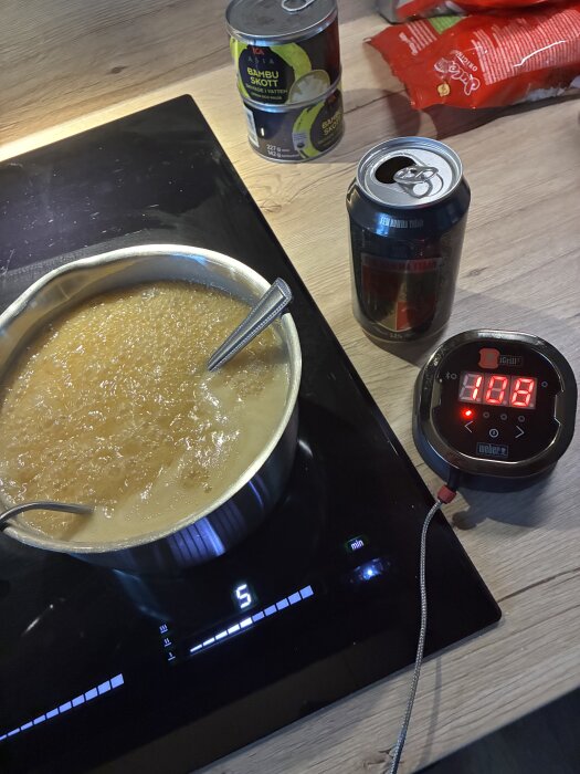 Kasansk gryta på spishäll, termometer visar 100°C, ölburk och bambuskottsburk på träbänk.