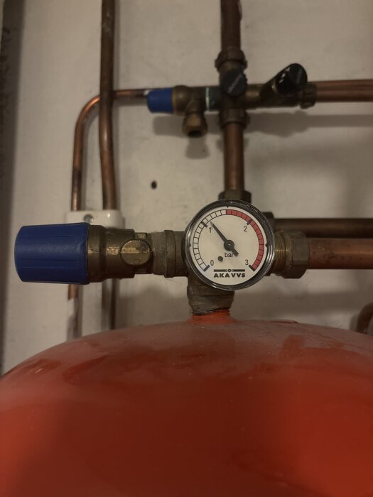 Tryckmätare på ett expansionskärl med kopparrör och blå ventil, visande ett tryck runt 1 bar, i en källare.
