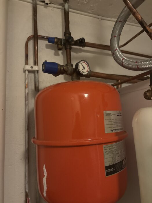 Rött expansionskärl i ett värmesystem med kopparrör, en tryckmätare och blåa ventiler synliga på väggen.