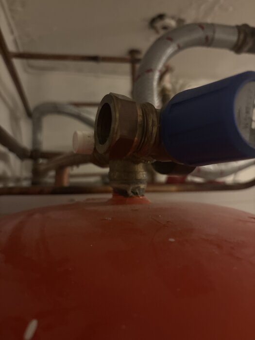 Närbild av en röd värmepumpsdel med anslutande rör och en blå ventil, samt en liten vit komponent i en källarinstallation.