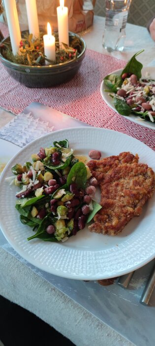 Sallad med spenat, sirapsstekt brysselkål, bönor, parmesan och schnitzel på en vit tallrik bredvid tända ljus på en röd duk.