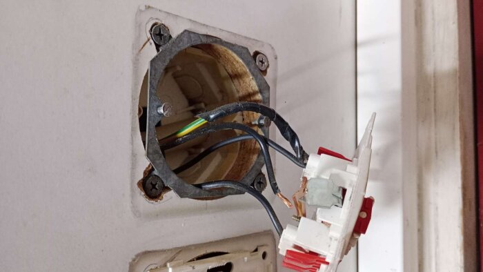 Öppen vägguttag med blottade elkablar, inklusive gröngul kabel tejpad svart, i ett äldre hus under elrenovering.