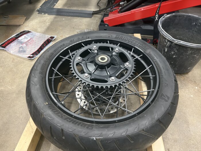 Motorcykeldäck med nytt JT-drev monterat. Finns i en verkstadsmiljö med verktyg och en pall i bakgrunden. Bridgestone Battlax Adventure däck.