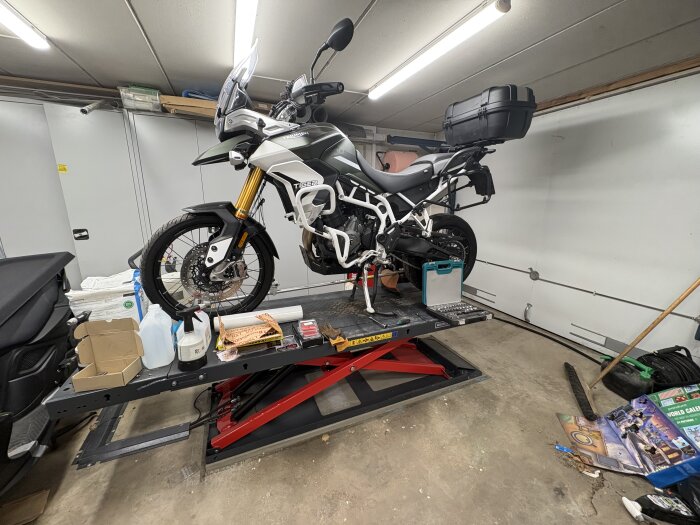 Motorcykel på lyft i garage med verktyg och produkter, omgiven av kartonger och rengöringsmedel.