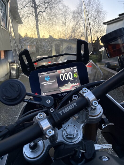 Stor närbild på Triumph Tiger-motorcykels instrumentpanel med nollhastighet och varning för låg temperatur. Bakgrunden visar en solnedgång.