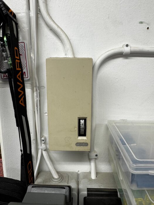 En termostat monterad på en vit vägg, omgiven av elektriska kablar och ett plastlock till höger, med en temperaturindikator.