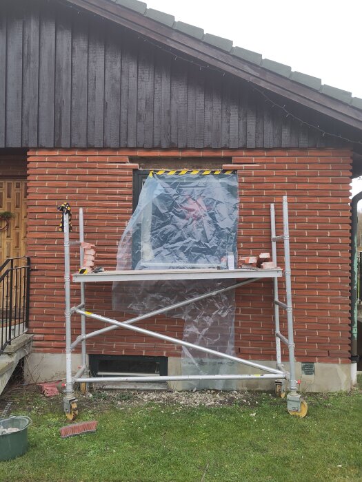 Byggställning framför tegelvägg med ett fönster delvis täckt av plast, armeringsarbete pågår.