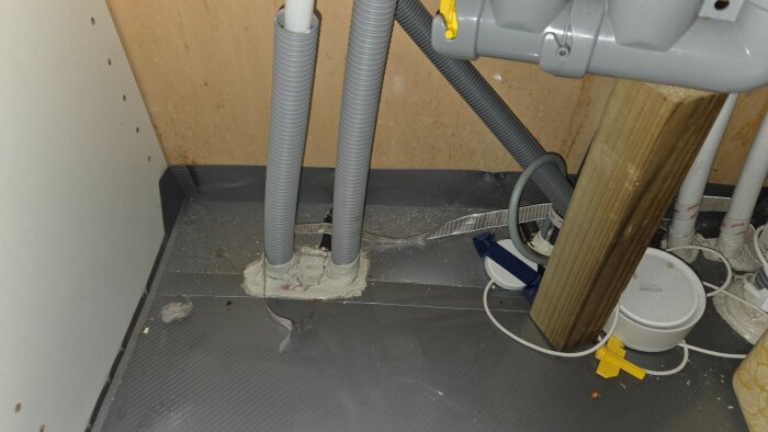 Stökigt avloppssystem under diskho med träregel 45x45 för att stabilisera, omgiven av rör och vattenlås i grått, sett på våt golvyta.