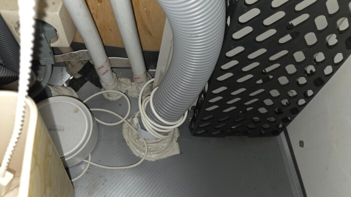 Avloppssystem under diskho med flera slangar och rör, inklusive en grå flexibel slang, omgiven av olika installationsdetaljer och kablar.