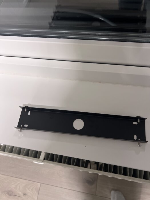 Svart fäste monterat på vit yta, visar installationsdetaljer med skruvar och hål. Bakgrund av fönster och golv.