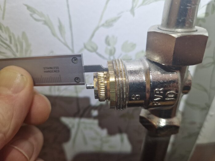 Närbild på en hand som mäter avståndet på en ventil med ett skjutmått i metall, intill ett rör med gänga.