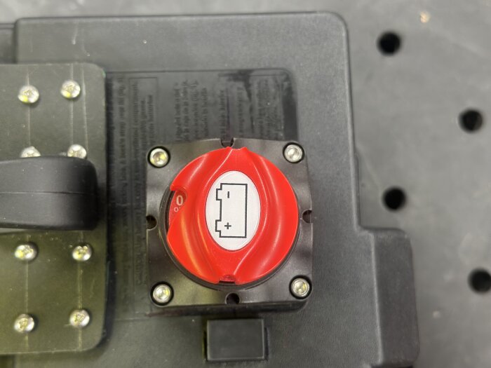 Röd huvudbrytare på en batterilåda med skruvar och handtag. Symbol för batterianslutningar synlig på knappen.