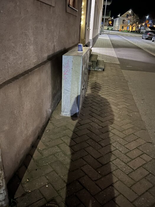 Skuggorna av en person sträcker sig över en trottoar vid en byggnad, en burk står på en elbox. Gatubelysning lyser upp kvarteret.