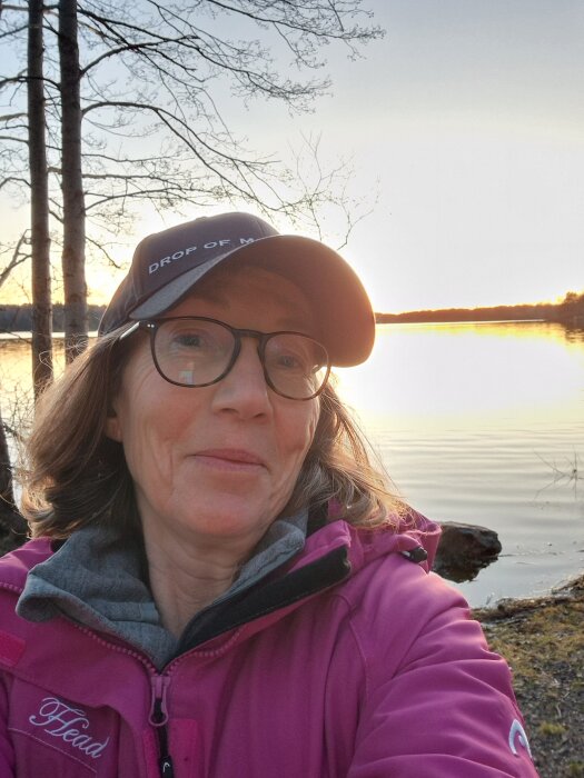 Person i lila jacka och keps vid sjö under solnedgång, träd i bakgrunden.