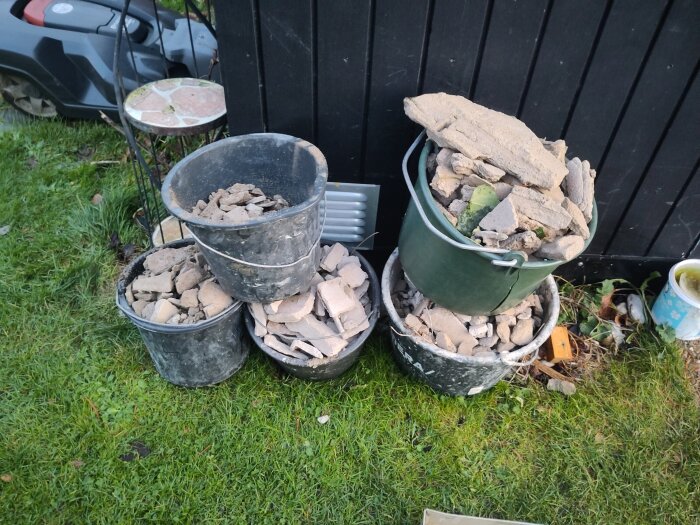 Hinkar fyllda med byggavfall och stenar står på en gräsmatta bredvid en mörk vägg. En robotgräsklippare syns i bakgrunden.