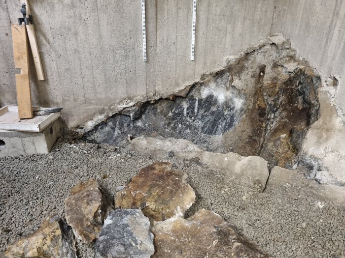 Bilden visar ett hörn av en betongvägg med lossad sten och grus på marken. Flera stora stenar ligger framför väggen efter att ha brutits loss.