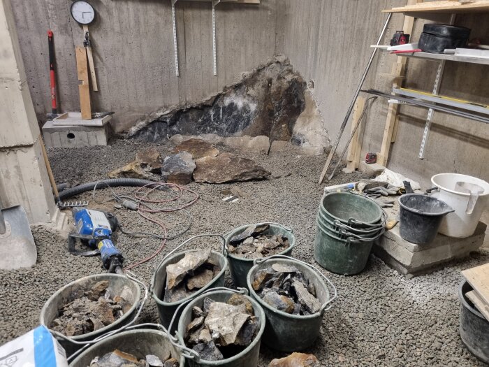 Hinkar fulla av sten, en borrmaskin på marken och en vägg av blottad berggrund i ett pågående byggprojekt.