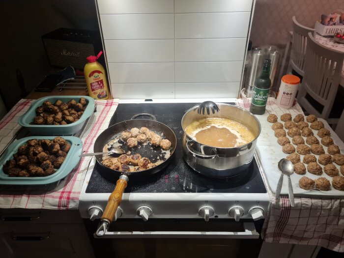 Stekpanna med köttbullar, såskastrull, två fyllda formar och en ugnsplåt med opreparerade köttbullar på spis med duk och redskap.