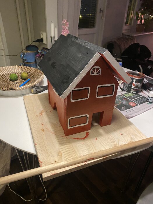 Rött fågelmathus med svart tak, designat som ett barndomshus, står på en träplatta i köksmiljö med verktyg och målarfärg i bakgrunden.