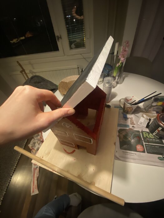 Hand håller upp taket på en miniatyr av ett rött hus som ska fungera som fågelmatare. Huset står på en träplatå med verktyg och målarfärger runtomkring.