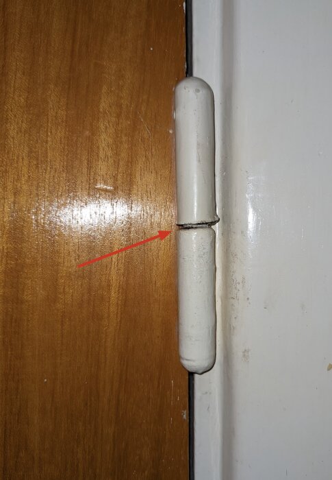 Övre gångjärnet på en brun dörr med en röd pil som pekar på en springa mellan gångjärnets delar.