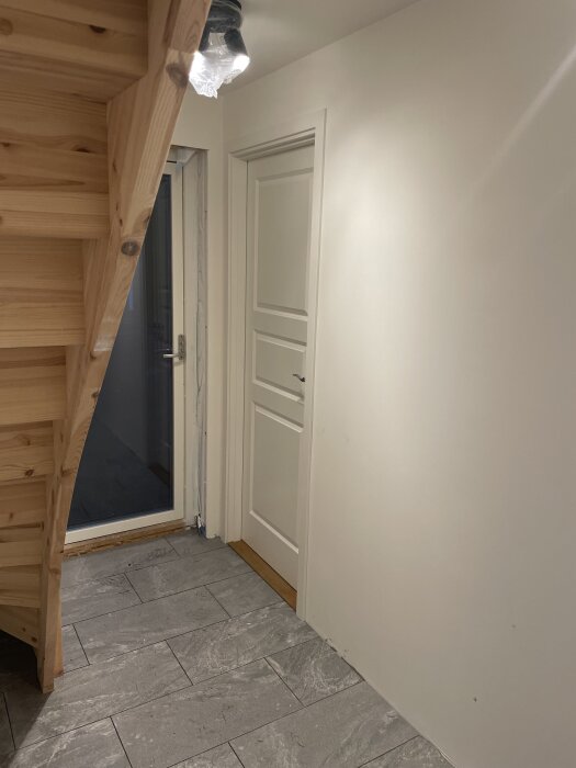 Trähylla i trä under trappa, ljus vägg, vit dörr och grått klinkergolv.