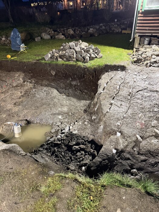 En byggplats med en grop fylld med vatten och stenrivor bredvid. Spruckna stenblock och en hög av stenar ses i gräset.
