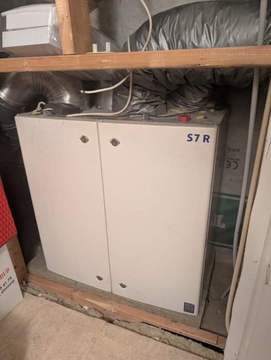Vit värmepumpsenhet med märkning "S7R" installerad i ett förråd med ventilationsrör och kablar.