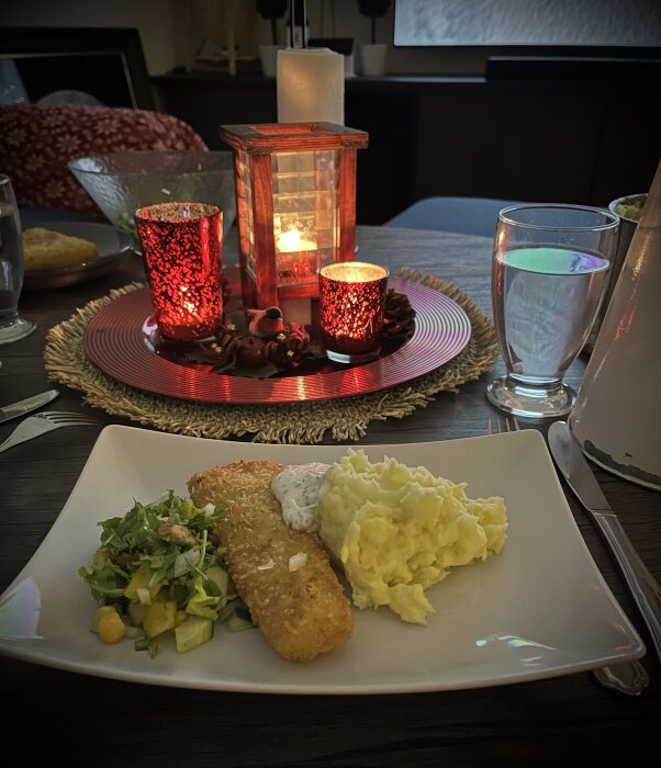 Panerad fisk med krispig yta, serverad med hemgjort potatismos, sallad och dillsås på en tallrik, omgiven av tända röda ljus.
