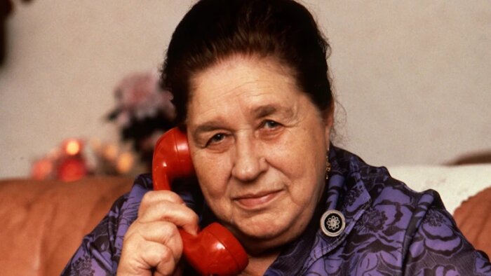 Kvinna i lila klänning håller en röd telefon mot sitt öra och ler, sittande i ett vardagsrum med dämpad bakgrundsdekoration.
