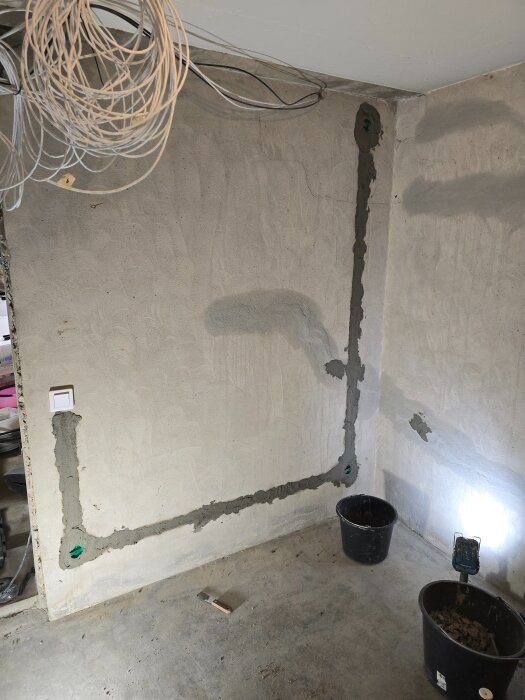 Betongvägg med tre eluttag installerade, synliga kablar och lagade områden runt hål och rör. Två svarta hinkar står på golvet.