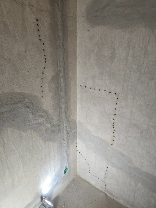 Hörn av betongvägg med borrhål för elinstallation, en ficklampa lyser upp området där väggdosor och flexrör ska installeras.