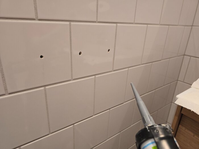 Vitt kakelvägg med två borrade hål, klar för montering av badrumstillbehör. Silikonverktyg syns i förgrunden.