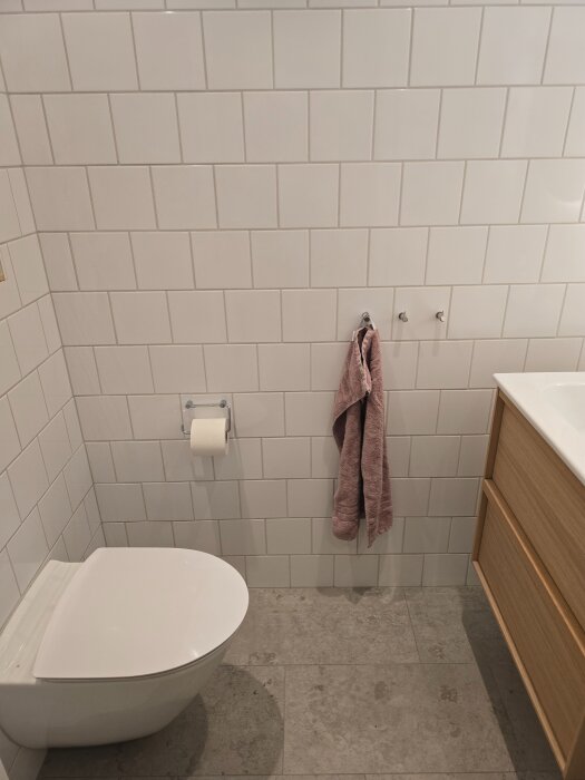 Badrum med vitkaklade väggar, toalettstol, handdukshängare med rosa handduk, toapappershållare och träskåp med vit tvättställ.