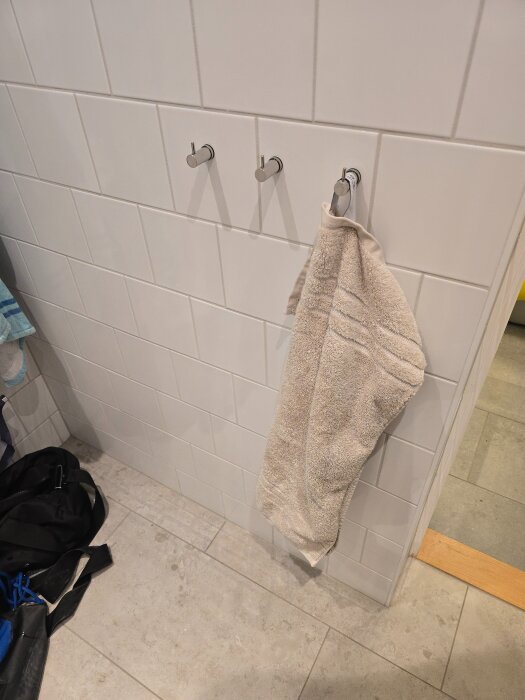 Vägg med klinkerplattor och tre monterade handdukskrokar, varav en krok har en beige handduk hängande.