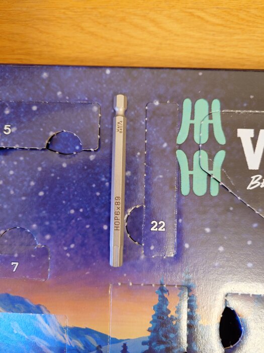 Borrbitten HOP 6x89 är placerad i en öppnad lucka nummer 22 på en verktygsadventskalender med vinterlandskap i bakgrunden.