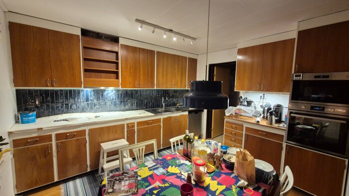 Kök under renovering med träskåp, mörk kakelvägg och verktyg på bänken. Matbordet är färgglatt uppdukat.