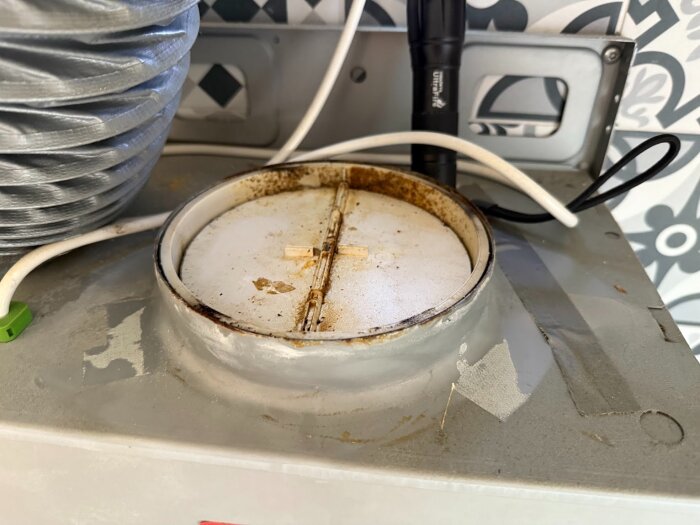 Spisfläktens spjäll med synliga fläckar och anslutna kablar. En ventilationsslang är kopplad bredvid. Spjället ser delvis demonterat ut.