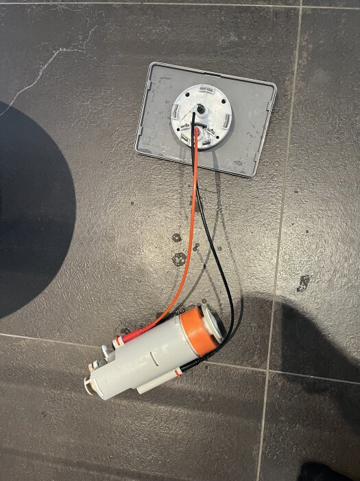 En toalettinsats ligger på ett klinkergolv, bestående av en grå cylinder med orange detaljer och kablar anslutna.