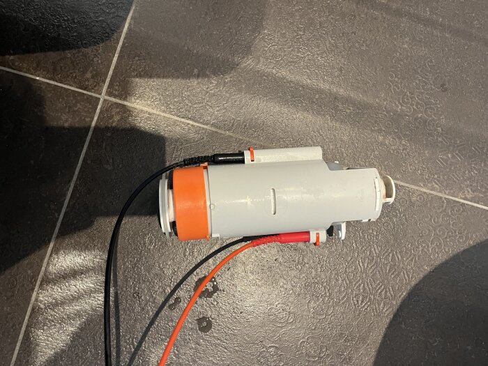 Komponent till toalett, grå plastcylinder med orange del, ligger på mörkt klinkergolv. Används för att identifiera nödvändig sits och insats.