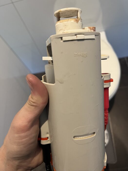 Insats till toalettspolning hålls i handen, vit plast med texten "triago" synlig, delvis nedsmutsad, del av toalettsits i bakgrunden.