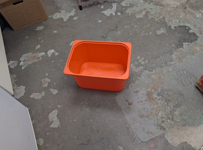 Orange behållare på golvet i ett garage, med några vattendroppar synliga i botten. Golvet visar tecken på fuktfläckar.