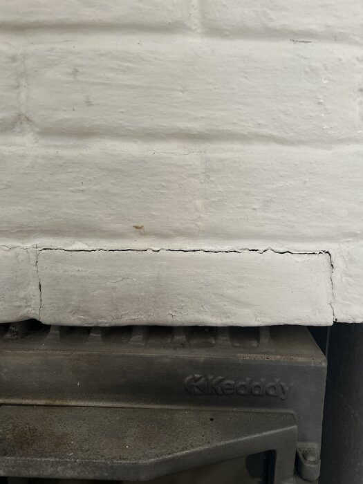Sprucken vitmålad tegelvägg med tegelsten som lutar mot en kamin. En spricka syns tydligt tvärsöver tegelväggen.