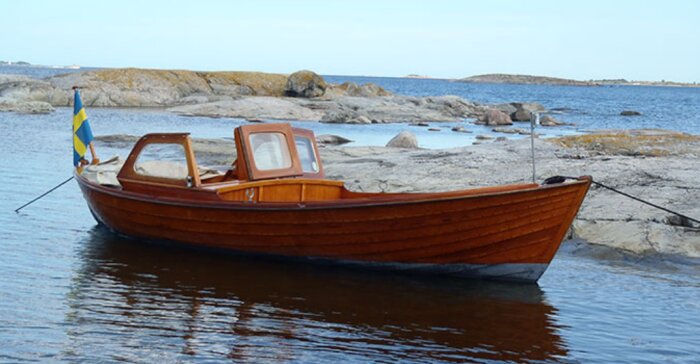 Träbåt med svensk flagga förankrad vid klippor i skärgårdsmiljö med blått hav och himmel i bakgrunden.
